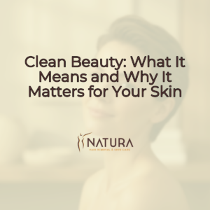 A square 1:1 image with a soft warm natural aesthetic. A tasteful female model with short hair and minimalist style, natural glowing skin, relaxed pose touching her face, and a genuine confident smile. The lighting is soft and diffused, and the background is slightly blurred. The image evokes a luxurious, natural, trustworthy, and spa-like mood, perfect for skincare promotion. The text "Clean Beauty: What It Means and Why It Matters for Your Skin" is overlaid in a modern sans-serif font, brown color, large size, and subtle shadow at the top-center. A minimalist "NATURA SPA" logo with a brand icon is at the center-lower-third.