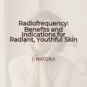 A woman with short hair and glowing skin, smiling warmly, touches her face. The background is softly blurred with warm beige and brown tones.